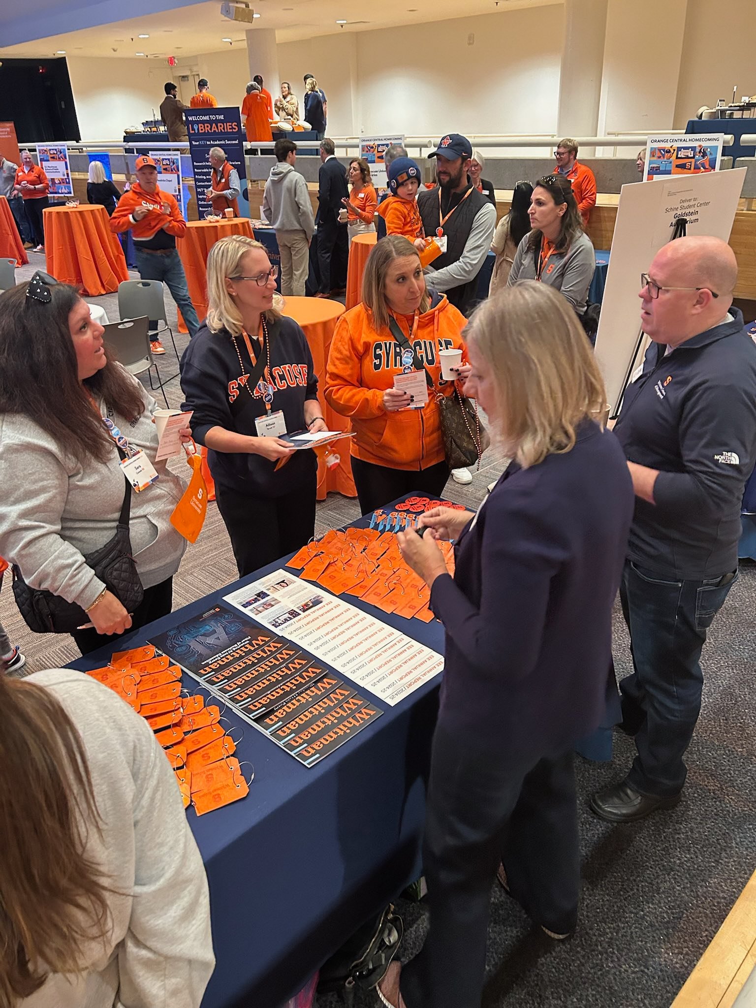 Alumni networking and conversing at indoor event with information tables displaying orange promotional materials and Syracuse bookmarks