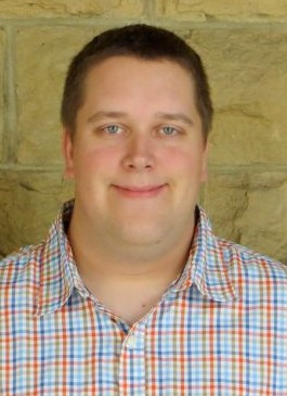 Person wearing a multicolored plaid button-up shirt standing in front of a beige stone wall.