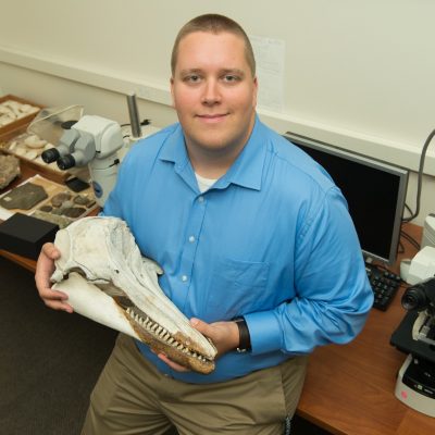 A person wearing a blue button-up shirt is seated in a laboratory setting, holding a large elongated skull with sharp teeth, likely from a marine mammal such as a dolphin or whale. The desk behind them contains various rock and fossil specimens, and there are two microscopes and a computer on the table, indicating a research or paleontology environment.
