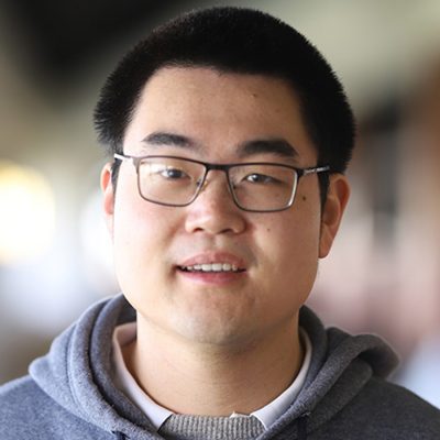 Professor Yizhi Liu, wearing black-framed glasses and a gray hoodie over a collared shirt, smiling at camera