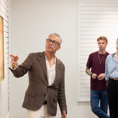 A person gestures toward a framed abstract painting with warm tones while three students observe in a white-walled gallery space