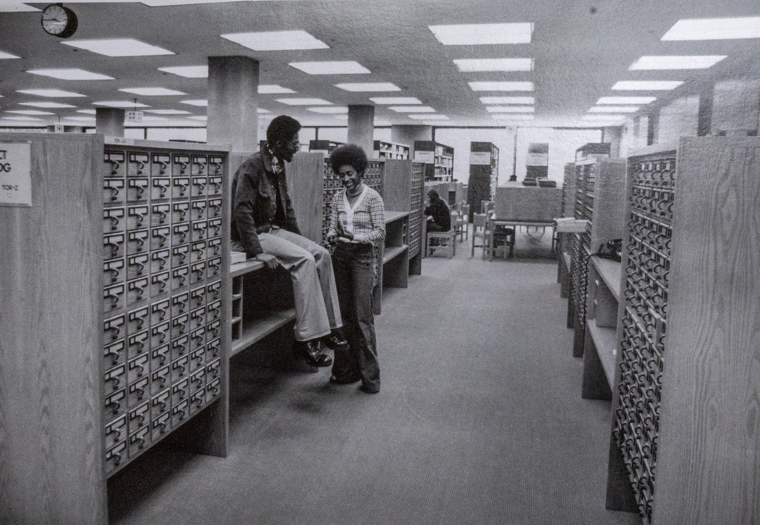 Black and white photo of a library card catalog area with two people interacting near wooden cabinets