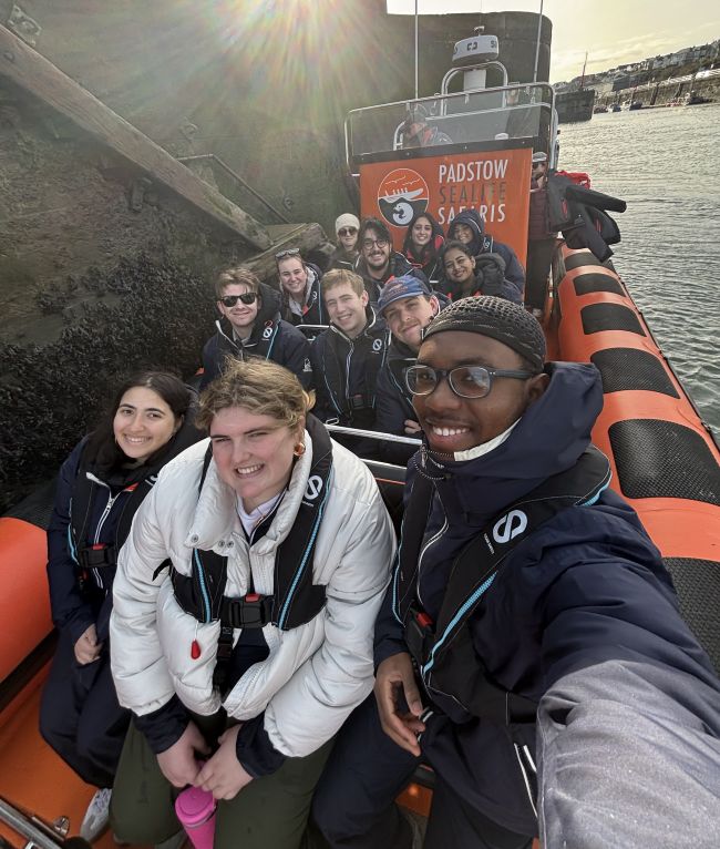 A group of students wearing life jackets smile for a selfie on a Padstow Sealife Safaris boat during sunset. A bright sun flare creates a dramatic effect in the sky as they prepare for their coastal excursion.