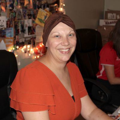 Person in orange top and brown head covering smiles at camera in cozy office with photo-covered wall and string lights.