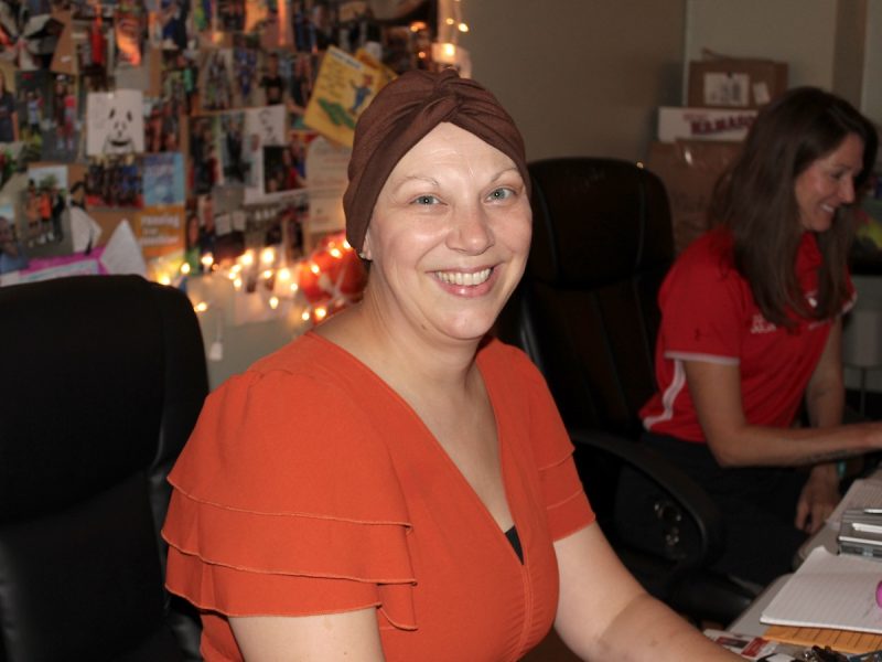 Person in orange top and brown head covering smiles at camera in cozy office with photo-covered wall and string lights.