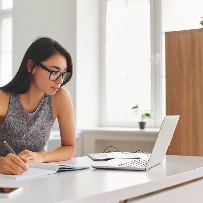 Person writing in notebook at desk with laptop and smartphone in bright office with plants and large windows