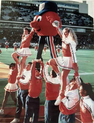Cheerleaders lift orange mascot in blue hat during stunt on football field, with stadium crowd in background.