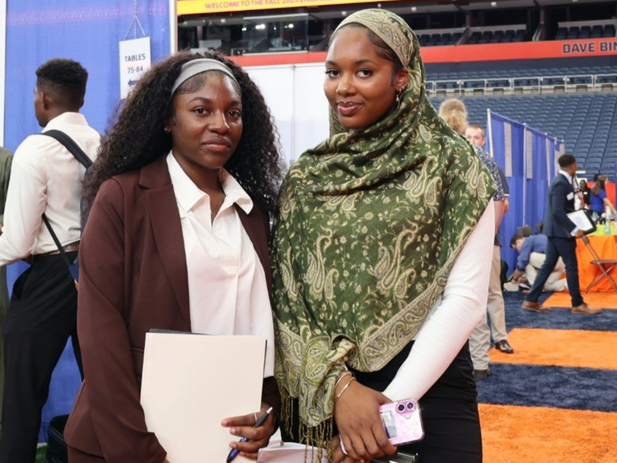 Two attendees at a Syracuse University career fair, standing in front of booths and signage in an indoor arena
