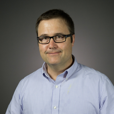 A person poses for a headshot in front of a grey backdrop.