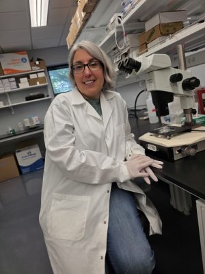 Person in white lab coat and safety glasses sitting at laboratory bench with microscope in background.