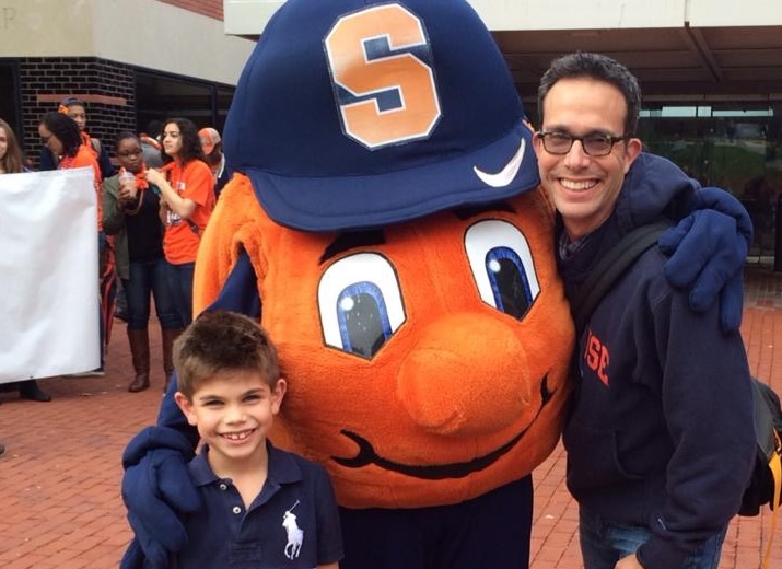 Child and adult posing with large orange mascot in blue 'S' cap, with crowd and building in background.