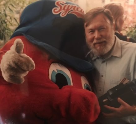 Person with camera stands beside orange mascot in 'Syracuse' hat giving thumbs-up.