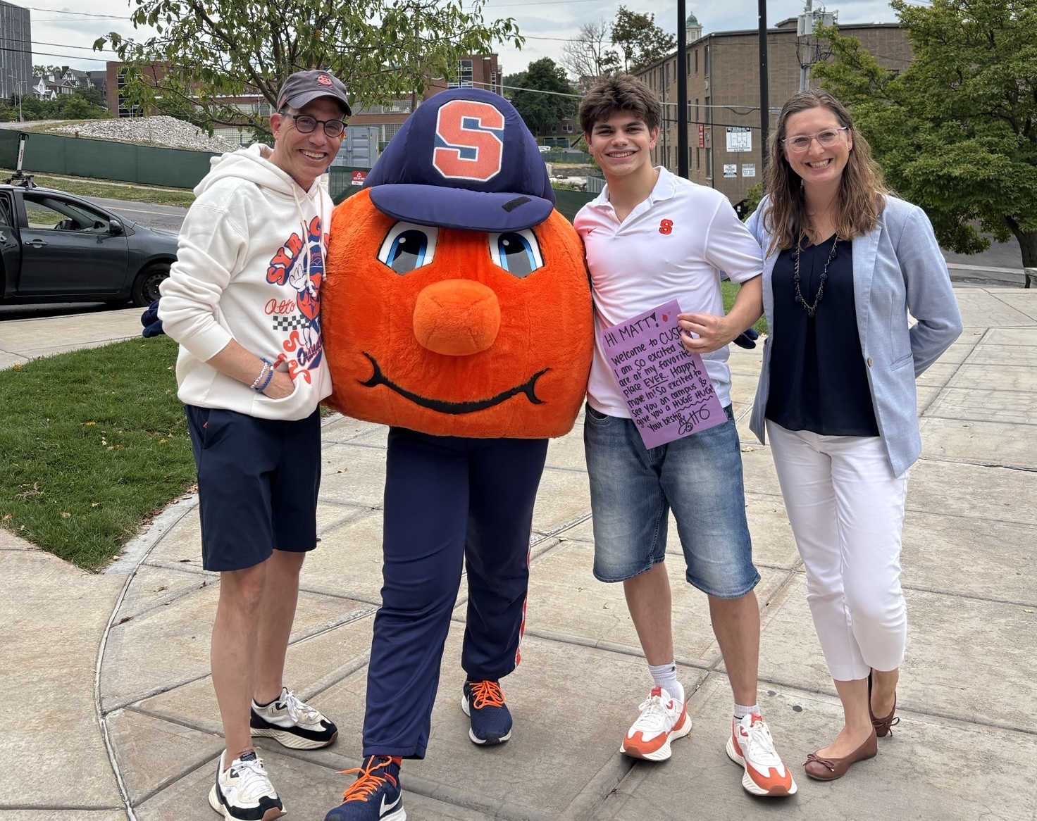 Four people, including orange mascot in 'S' cap, pose on sidewalk with trees and buildings in background.