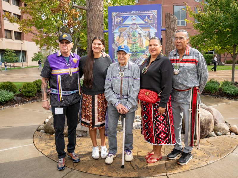 Five people stand together in front of a decorative panel featuring Indigenous artwork on campus