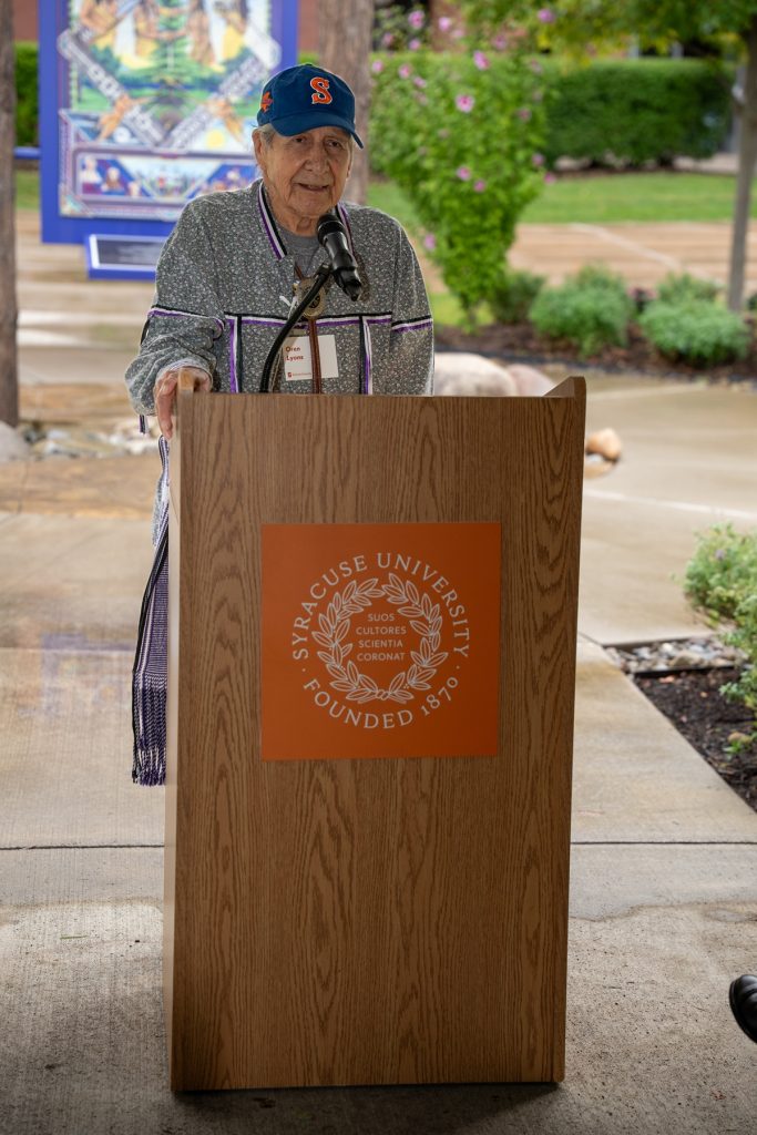 A person speaks at podium wearing traditional Indigenous attire and Syracuse cap