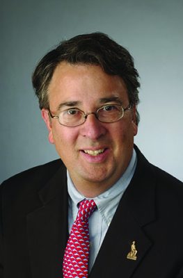 Person wearing a suit and tie, against a gray background.