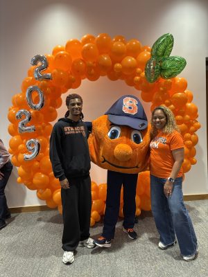 Two people pose with orange mascot under balloon arch with '2029' in silver.