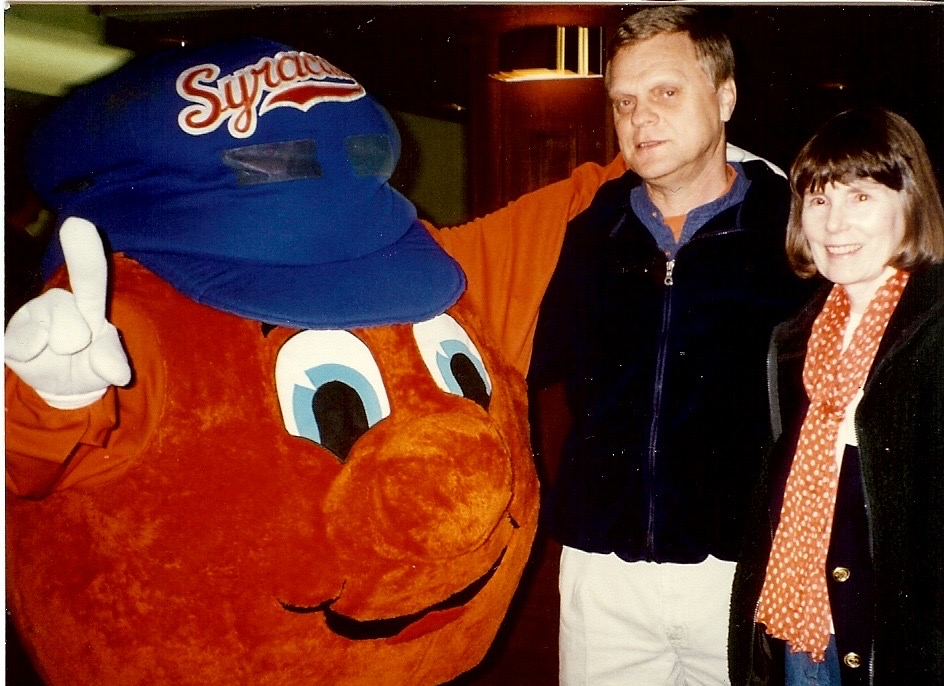 Two people pose with Syracuse orange mascot in blue 'Syracuse' hat.