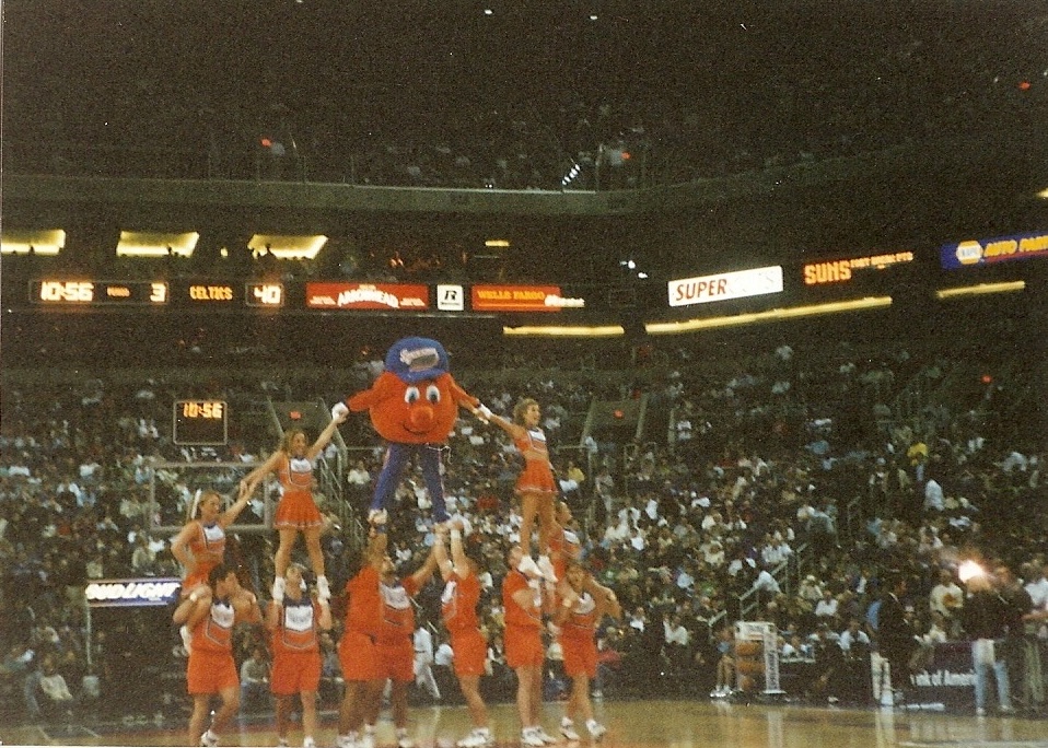 Cheerleaders perform stunt with orange mascot atop formation on basketball court, crowd and scoreboard in background.