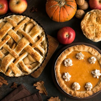 Assorted autumn pies on a dark surface with apples, pumpkins, spices, and nuts.