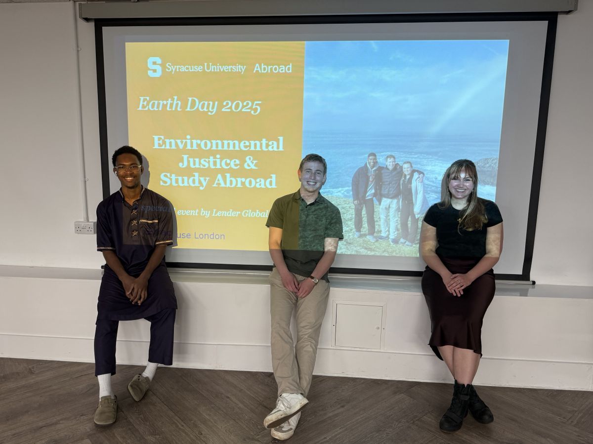 Three people stand in front of a projection screen displaying "Earth Day 2025: Environmental Justice & Study Abroad" with the Syracuse University Abroad logo. The presentation is an event by Lender Global and Syracuse London, with a coastal photo visible on the right side of the screen.