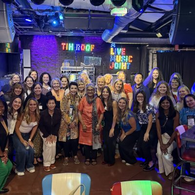 Group photo of several people gathered in a colorful indoor venue with stage lighting, exposed ceiling pipes, and neon signs in the background.