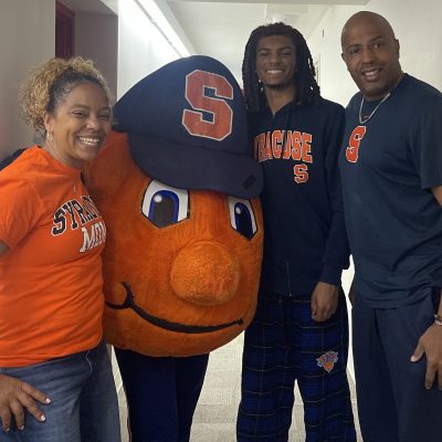 Three people in Syracuse University gear pose with Otto the Orange mascot in a hallway.