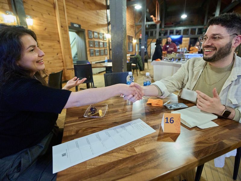 Two people shake hands and smile across a wooden table in a room with others conversing in the background.
