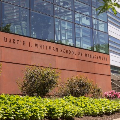Exterior of the Martin J. Whitman School of Management building with large glass windows, red-brown wall, and landscaped greenery and flowers in front.