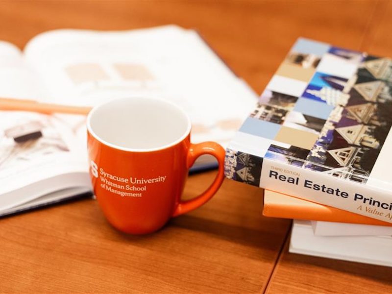 Orange Syracuse University Whitman School mug on a wooden table beside an open book, stack of textbooks, and orange pen.