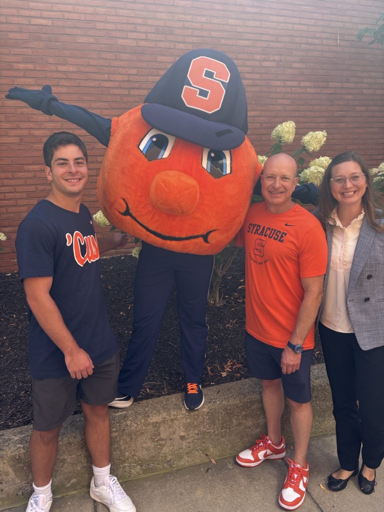 Group photo with Otto the Orange mascot and Syracuse University supporters posing in front of a brick wall.