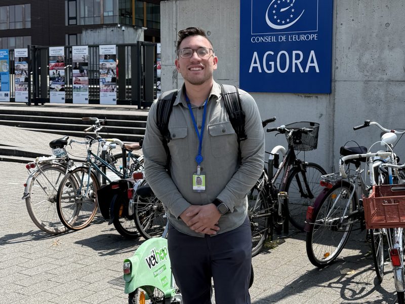 Person standing outside the Council of Europe Agora building, wearing a gray shirt with a lanyard and backpack, with bicycles parked nearby and informational posters visible in the background