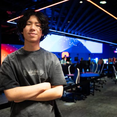 Person standing with arms crossed in an esports venue, featuring rows of gaming chairs and large illuminated screens in the background.