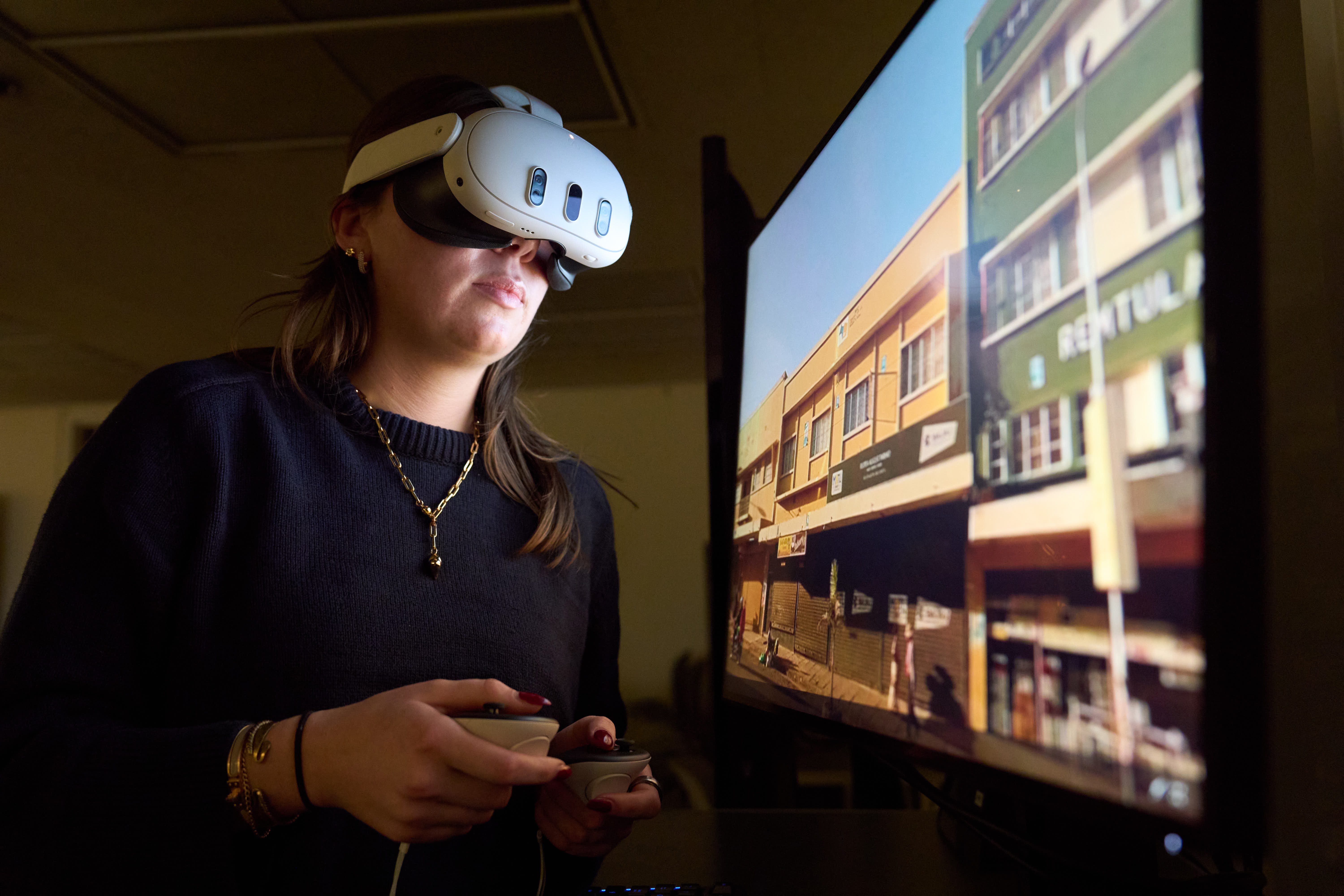Person wearing a VR headset and holding controllers while standing in front of a monitor displaying a street scene with buildings and signage.