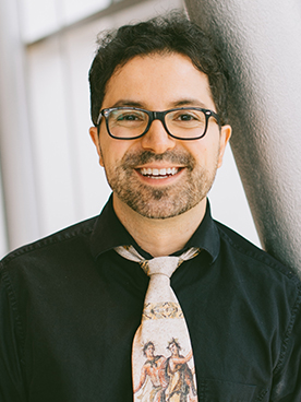 A professional headshot of Kivanc Avrenli, a Syracuse University professor, wearing glasses, a black collared shirt, and a distinctive illustrated tie. He is smiling at the camera while leaning against a white wall.