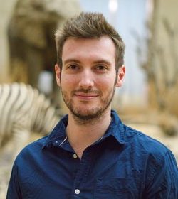 Portrait of a young person in a blue patterned shirt smiling at the camera, with museum exhibits in the background.