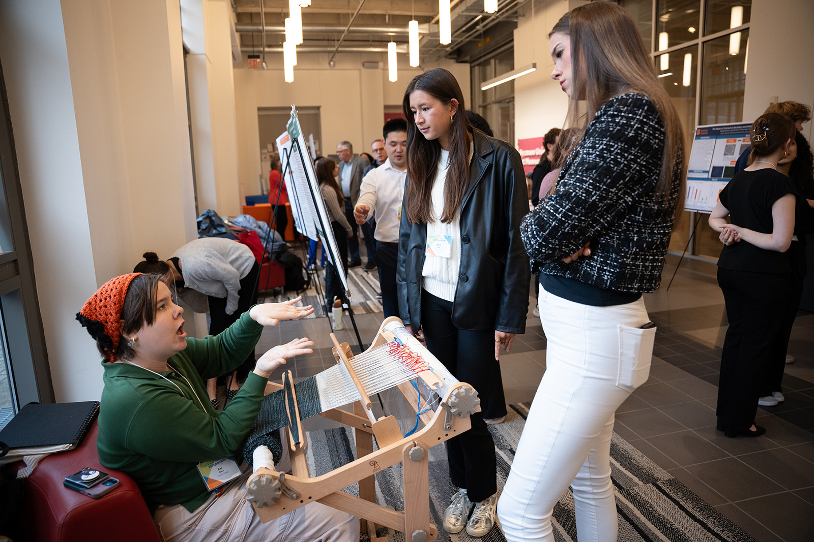 Person seated at a small loom demonstrating weaving techniques to two onlookers in a busy indoor event space with posters and attendees in the background