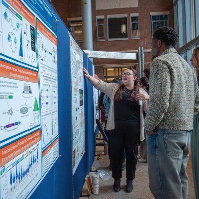 A student in glasses points to a research poster while explaining her work to two other students at a poster presentation session