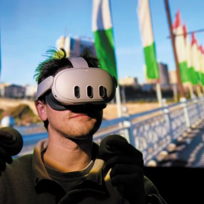 Person wearing a VR headset and holding two controllers, with a large screen in the background displaying a row of green-and-white flags along a waterfront promenade.