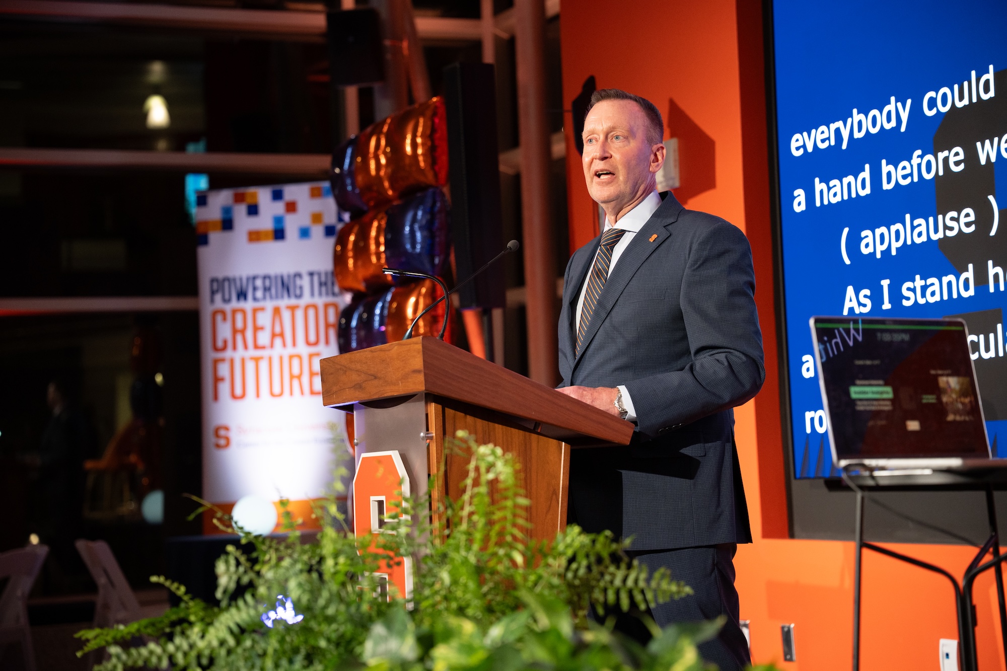 Person standing at a podium with a microphone, wearing a dark suit and striped tie. Behind the podium are orange and blue balloons and a banner reading “Powering the Creator Future.” A large screen with text is visible to the right.