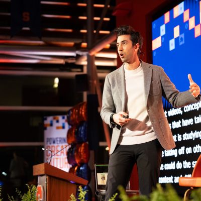 Person giving a presentation on stage, wearing a light gray blazer and white sweater, holding a remote control. A large screen with text and colorful graphics is behind them, and a podium with balloons is visible to the side.