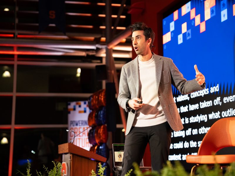 Person giving a presentation on stage, wearing a light gray blazer and white sweater, holding a remote control. A large screen with text and colorful graphics is behind them, and a podium with balloons is visible to the side.