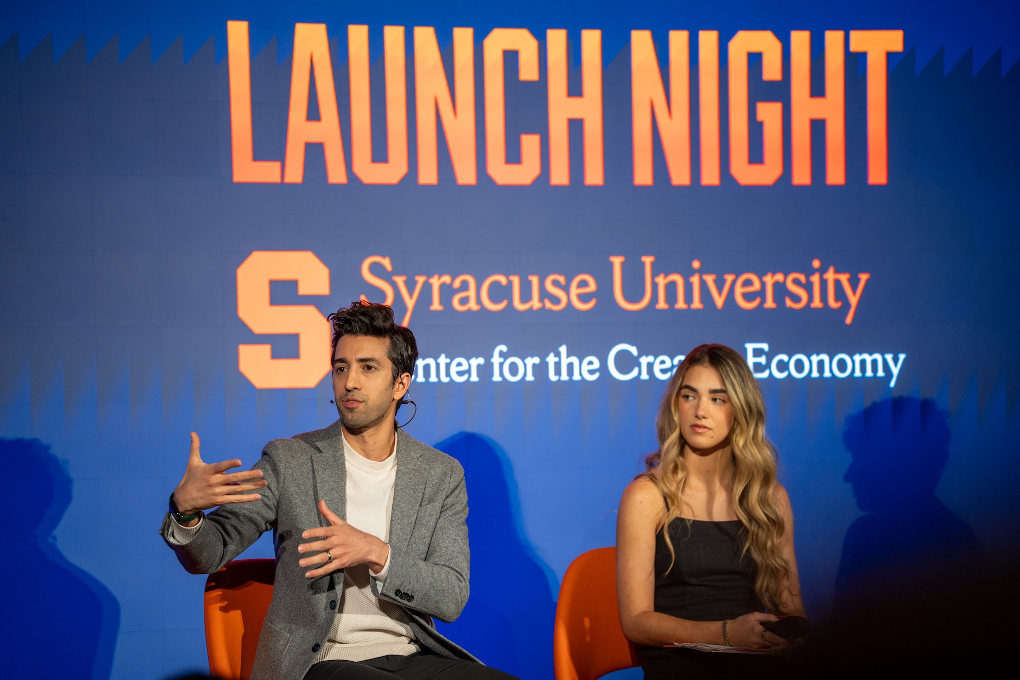 Two people seated on stage in front of a large screen displaying “Launch Night” and “Syracuse University Center for the Creative Economy.”