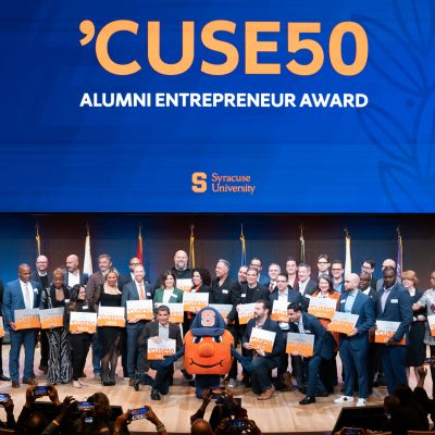 Large group of 'CUSE50 Alumni Entrepreneur Award recipients posing on stage with Otto the Orange mascot, holding orange award signs beneath Syracuse University branding and international flags.