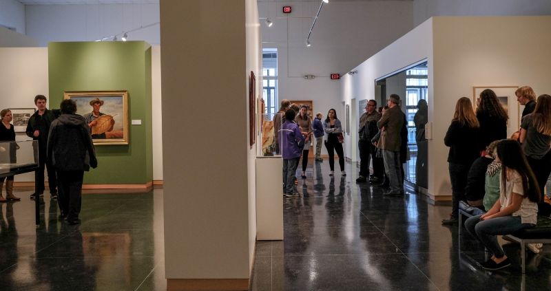 Visitors explore gallery spaces at an art museum, viewing paintings and sculptures displayed in rooms with colorful accent walls, track lighting and polished concrete floors.