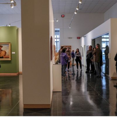 Visitors explore gallery spaces at an art museum, viewing paintings and sculptures displayed in rooms with colorful accent walls, track lighting and polished concrete floors.