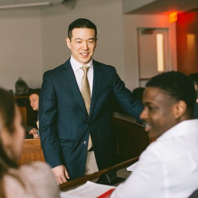 Professor in a suit and tie stands in front of three students who are seated