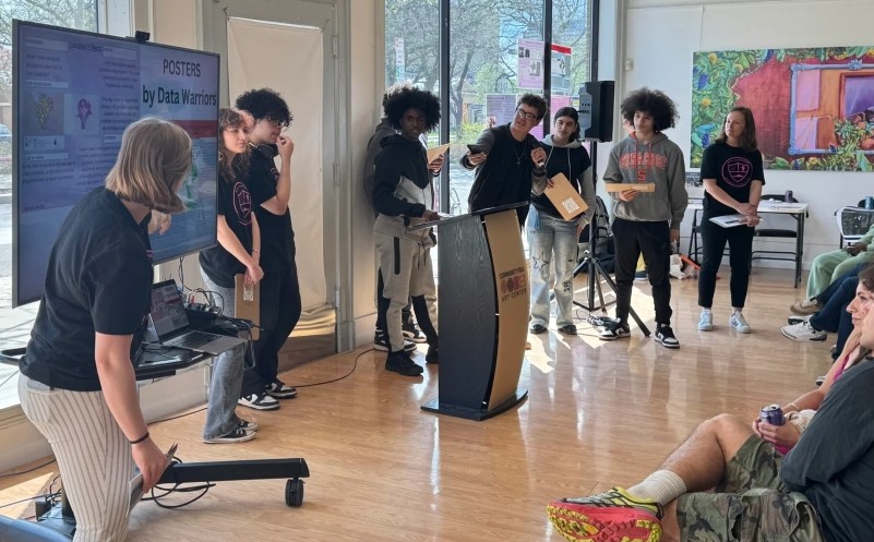 A group of students in matching black t-shirts present their work around a "POSTERS by Data Warriors" display board in a bright room with hardwood floors and large windows.