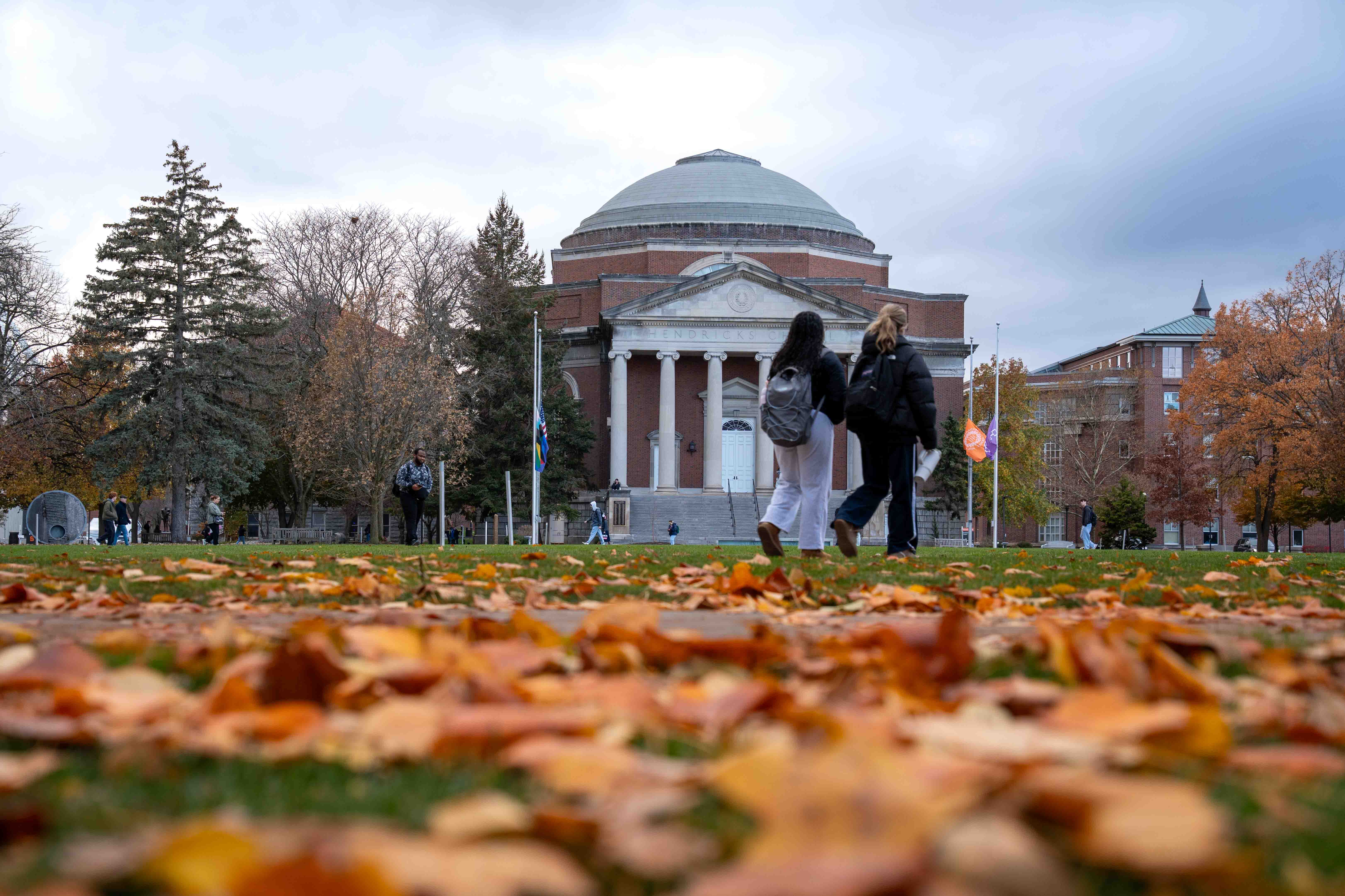 Historic domed building with columns viewed across a lawn covered in autumn leaves, with two people walking in the foreground and trees showing fall colors