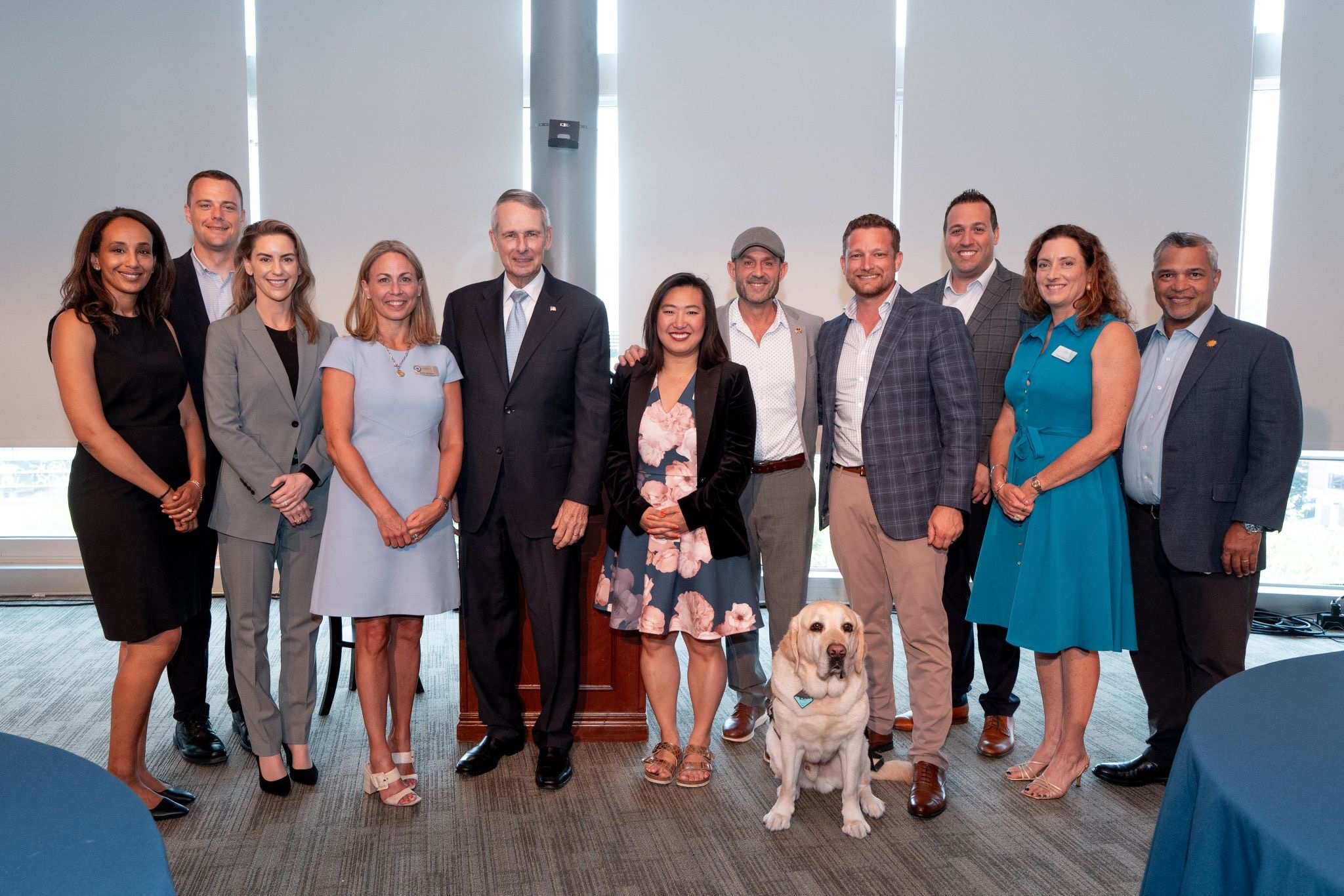 Group of people standing together indoors in front of large windows, with a yellow Labrador sitting in front of them.